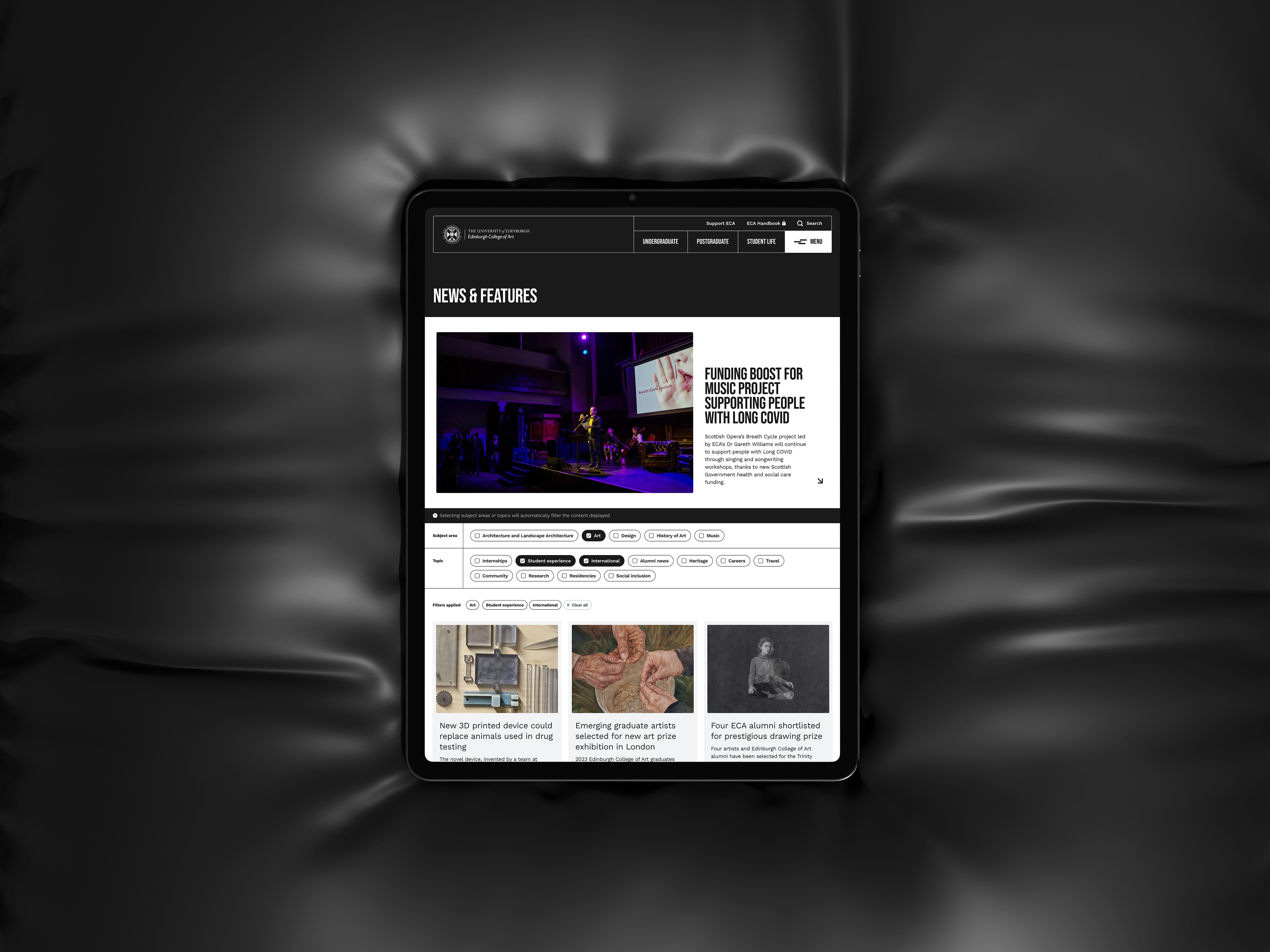 Tablet on a dark surface displaying the Edinburgh College of Art website news section with articles and features.