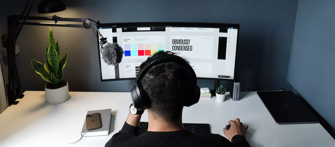 The image shows a guy sitting at a desk, looking at the computer screen. On the screen it's possible to see some colour palettes, a white picture with "Obviously condensed" in black capital letters written on it, and other digital elements.
