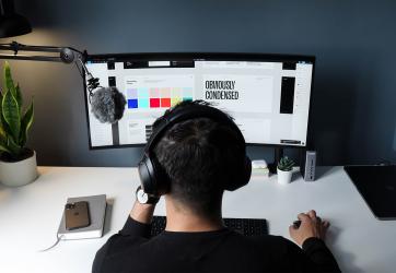 The image shows a guy sitting at a desk, looking at the computer screen. On the screen it's possible to see some colour palettes, a white picture with "Obviously condensed" in black capital letters written on it, and other digital elements.