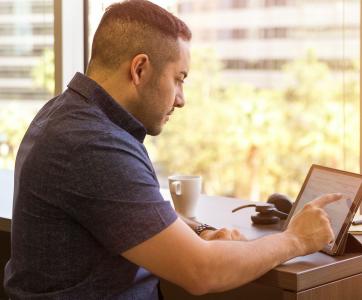 Man sitting at a high table in front of a window on a touch screen tablet device.