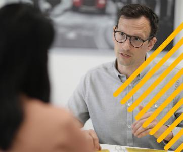 Man in glasses and gray shirt talks while gesturing to a woman across from him in an office setting.