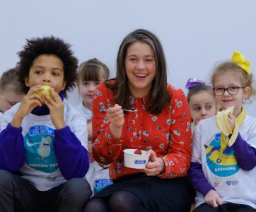 School children and a teacher sit in a row, eating fruit and smiling