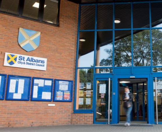 Entrance of St Albans City & District Council building with blue-framed glass doors, noticeboards, and the council crest on a brick wall