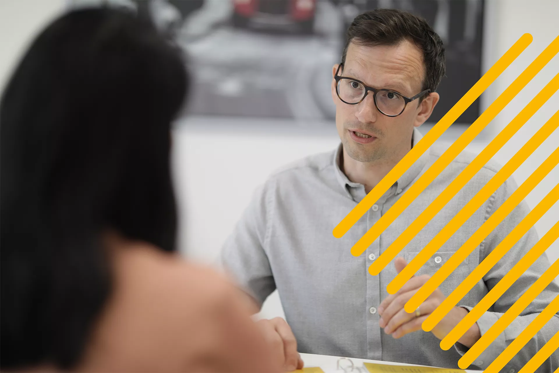 Man in glasses and gray shirt talks while gesturing to a woman across from him in an office setting.